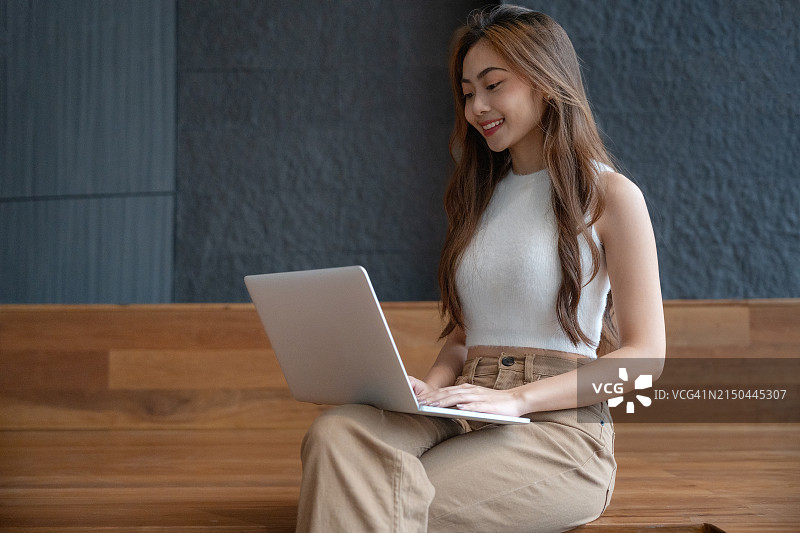 快乐的小女孩在公园里用笔记本电脑工作。年轻的亚洲女人坐在户外咖啡馆里参加在线会议。对着笔记本电脑摄像头说话。解释一些东西。喝咖啡。图片素材