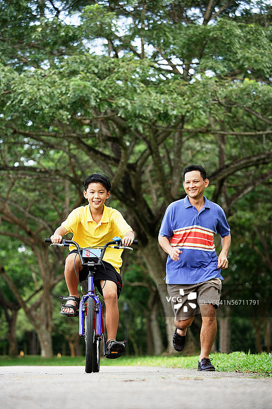 父子在公园里，儿子骑自行车，父亲在旁边跑步图片素材
