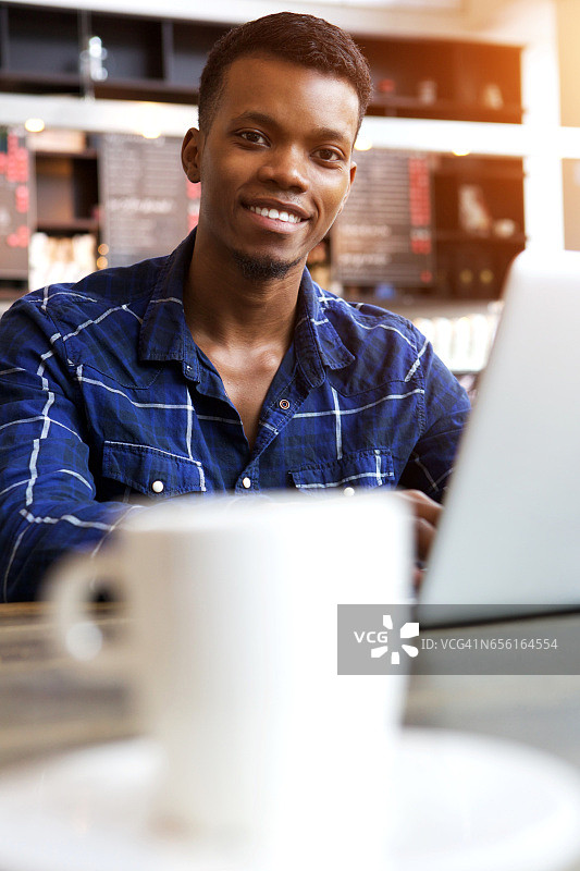 微笑的年轻人坐在咖啡馆，拿着一杯咖啡和笔记本电脑图片素材