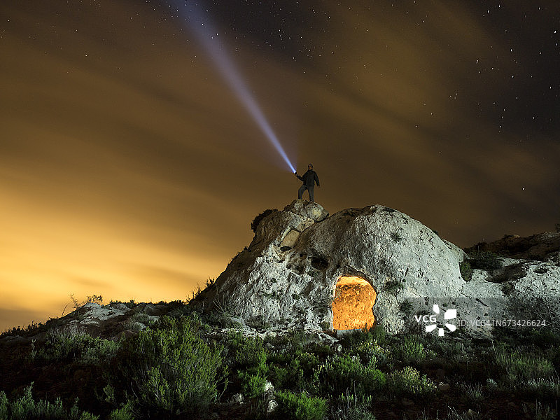 山顶上有一个古老的避难所，在岩石中挖掘，一个夜晚的天空与橙色的云和星星，与一个人做发光的信号与灯笼图片素材
