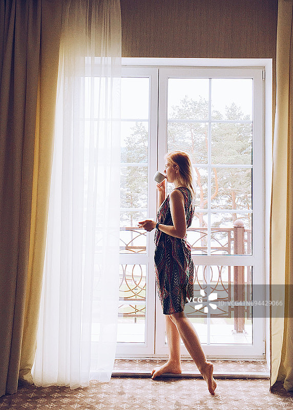 年轻的金发女子站在窗边，用杯子喝咖啡或茶。图片素材