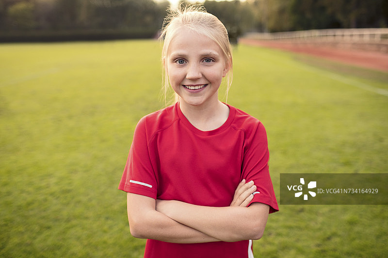 穿着红色t恤的快乐女孩的肖像，双手交叉站在足球场上图片素材