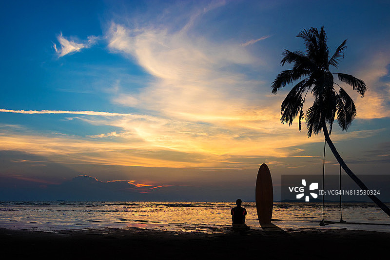 冲浪男子的剪影坐在海滩上冲浪板。冲浪场景在日落海滩与彩色的天空。户外水上运动冒险生活方式。夏天的活动。20多岁英俊的亚洲男模。图片素材