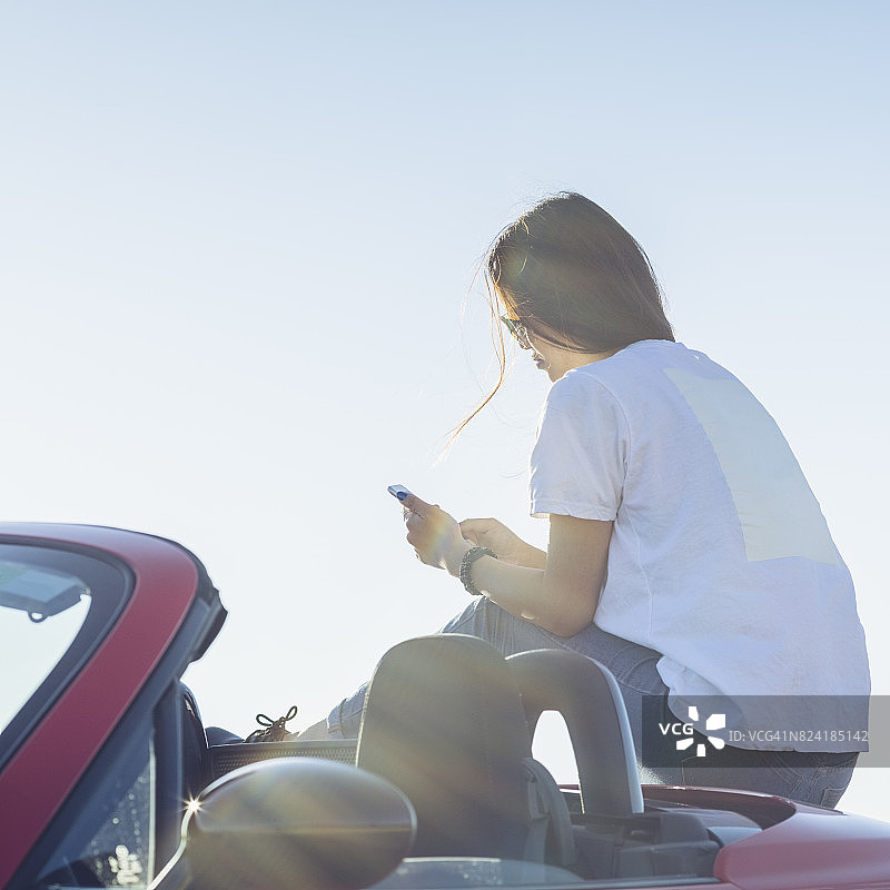 年轻的亚洲女人坐在红色敞篷车上，看着智能手机，图片素材