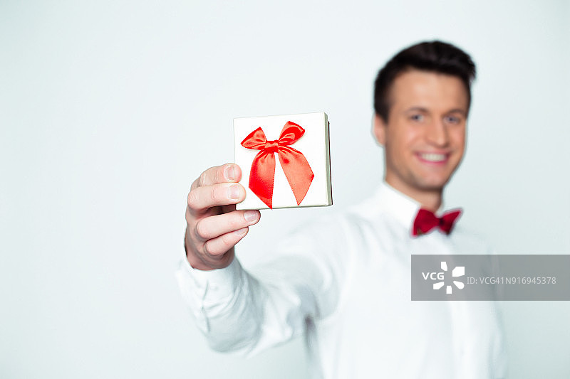 给心爱的人的礼物。年轻美丽的微笑优雅的男人在白色衬衫和红色领结拿着一个盒子与礼物，并显示它的相机。情人节。图片素材