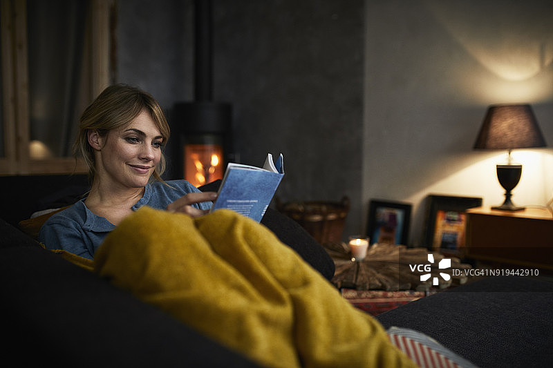 微笑的女人晚上在家沙发上看书的肖像图片素材