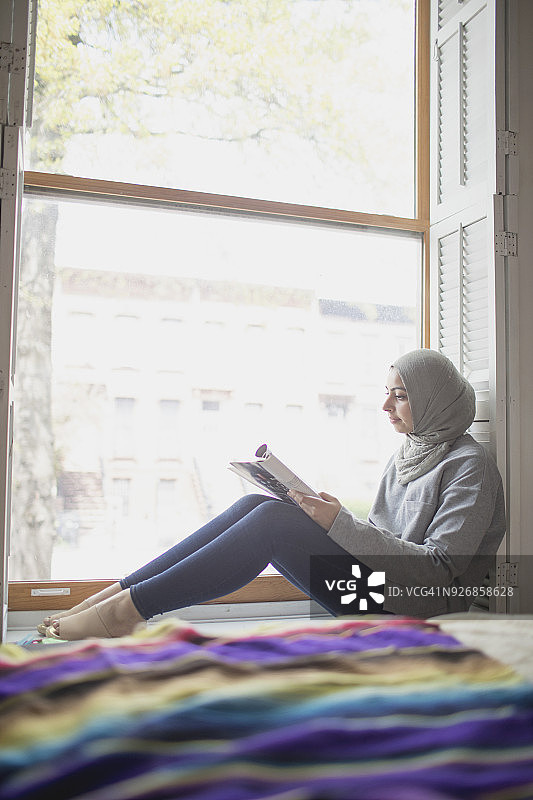 一个穆斯林妇女戴着头巾，在窗台上阅读图片素材