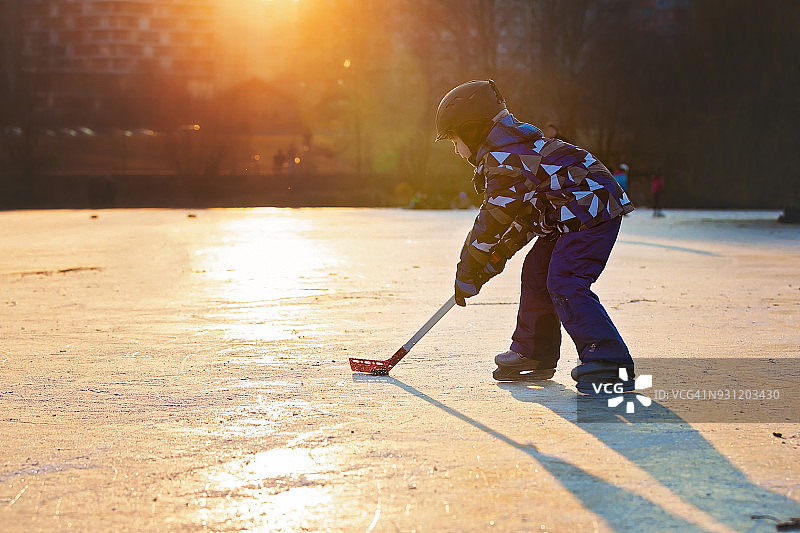 孩子们在公园里冰封的湖面上打曲棍球和滑冰，冬天的日落时分图片素材