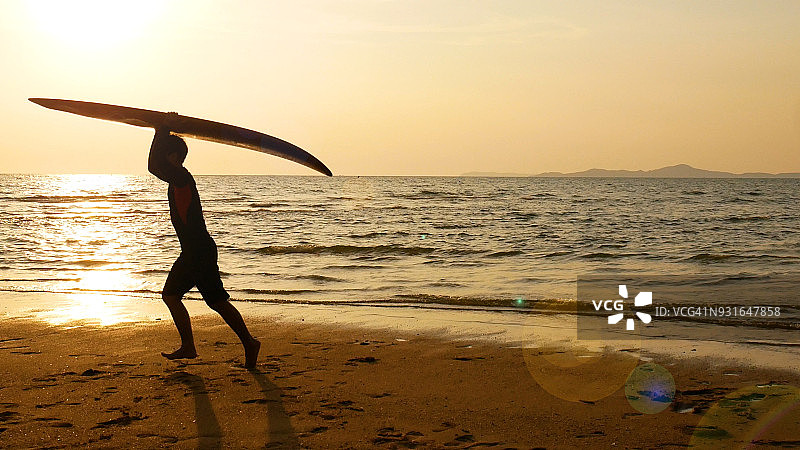 在热带海滩日落时，快乐的冲浪人跑着长冲浪板的剪影。在美丽的日落时分，在海边的海滩冲浪者。冲浪水上运动户外活动图片素材