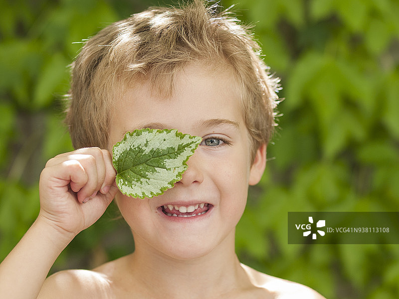 夏日里，小男孩微笑着，用一片斑驳的叶子遮住一只眼睛图片素材