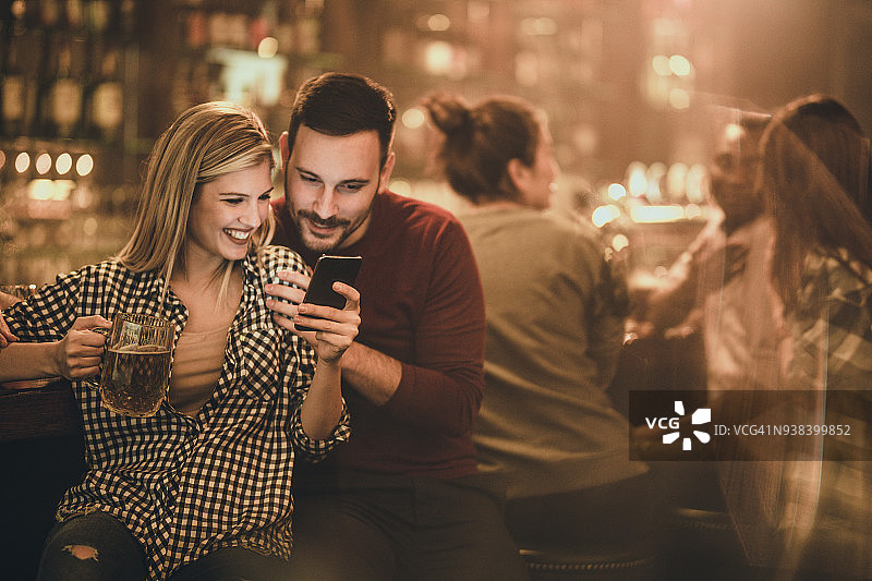 一对年轻幸福的情侣在酒吧过夜时，正在看智能手机上的短信。图片素材