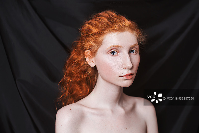 在黑色的背景下，长长的红卷发扎成马尾辫。红发女孩，白皙的皮肤，蓝眼睛，不化妆的样子很不寻常。自然美景。文艺复兴时期的女孩图片素材