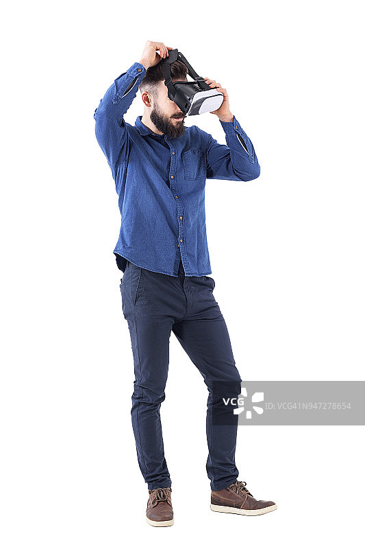 年轻的大胡子男子的侧面视图试着戴上虚拟现实头盔向下看。图片素材