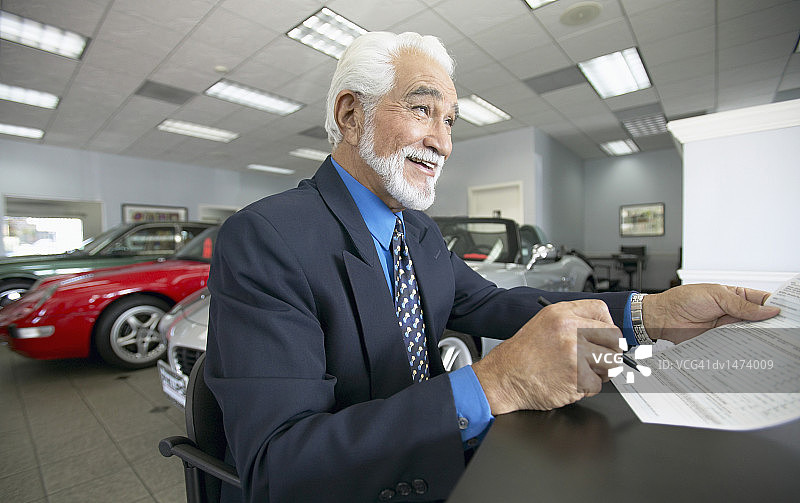 高级销售人员手持文件坐在汽车展示厅的一张桌子前图片素材