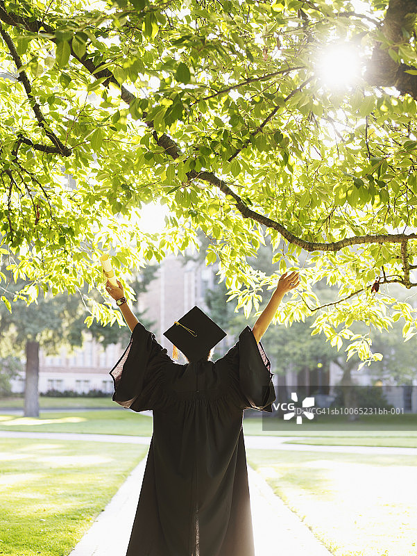 年轻女子在毕业礼服与举起的胳膊户外，后视图图片素材