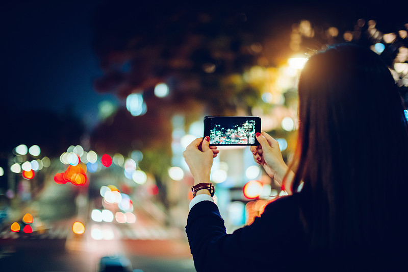 一名女性用智能手机拍摄东京繁忙城市夜景的后视图图片素材