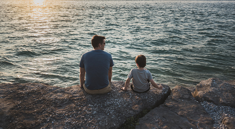 父亲和儿子坐在海边的岩石在日落的背影图片下载