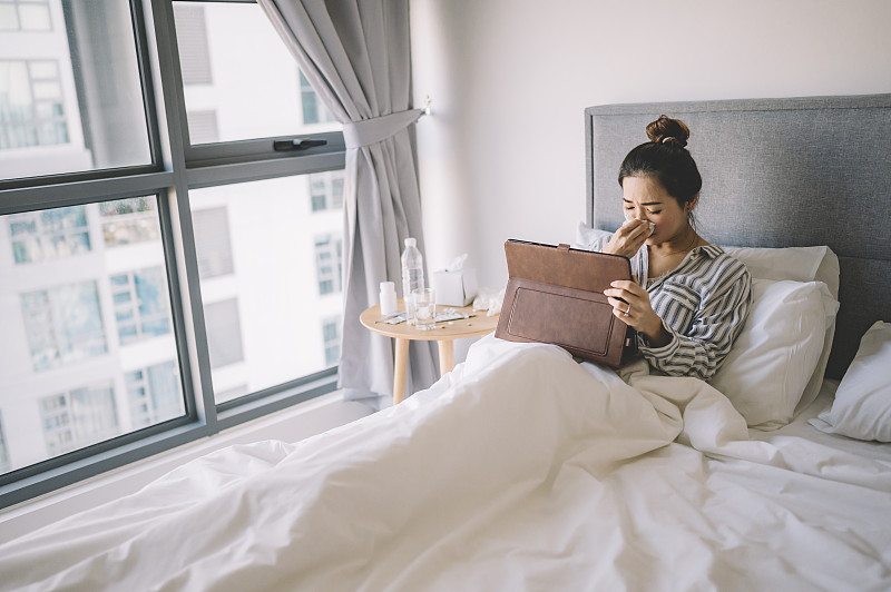一位亚洲华裔女性在床上穿着睡衣，用纸巾捂着鼻子看数字平板电脑图片下载