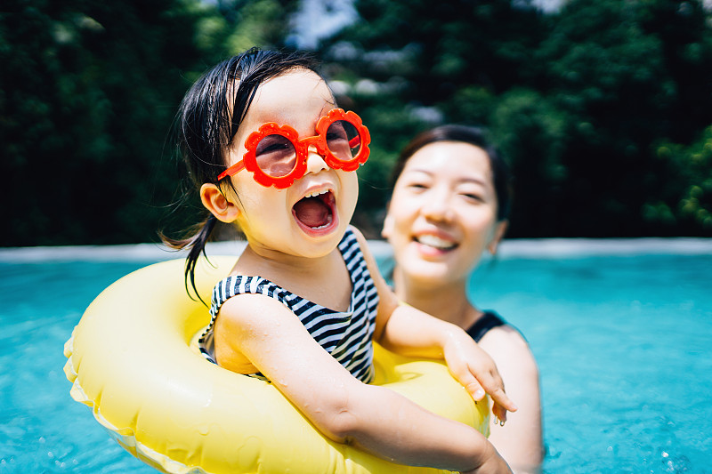 快乐的亚洲蹒跚学步的女孩与太阳眼镜愉快地微笑，享受家庭团聚的时间与母亲有乐趣在游泳池在夏天图片下载