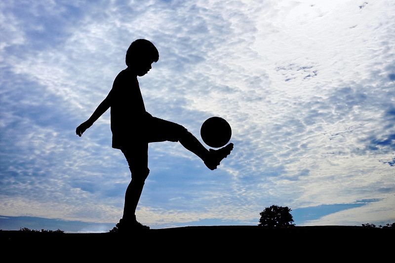 日落时分，在多云的天空下，一个男孩在球场上踢足球图片下载