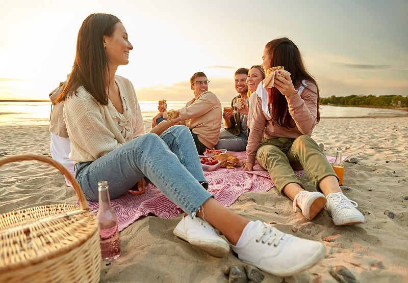 友谊，休闲和快餐概念-一群快乐的朋友吃三明治或汉堡在海滩野餐在夏天。快乐的朋友在海滩上野餐吃三明治图片素材