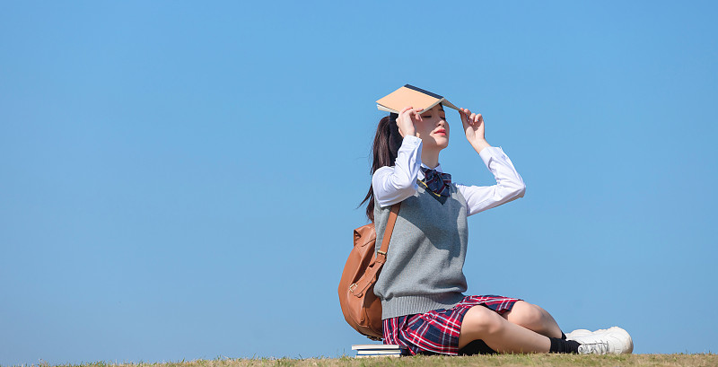 蓝色天空背景下，坐在草地上的亚洲女学生图片下载