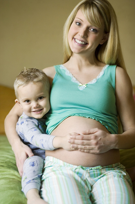 年轻的孕妇抱着儿子(4-5岁)摸着肚子躺在床上，画像图片下载