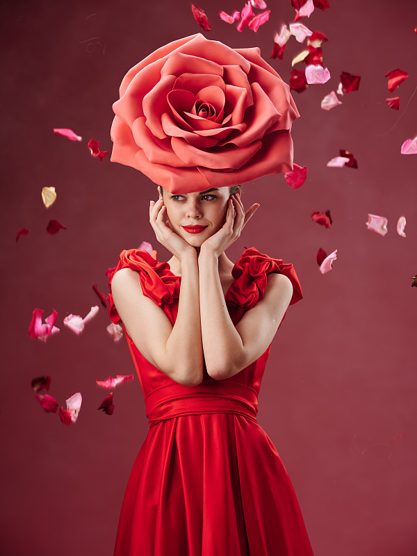 一个有着完美五官和美丽身材的年轻女孩，穿着合身的红色衣服，站在她的头上红色的背景上，戴着一顶玫瑰形状的帽子。图片下载