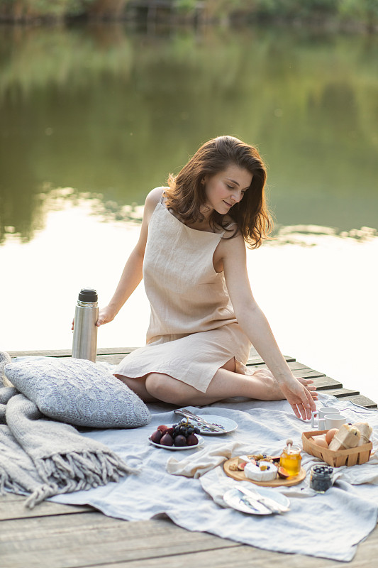 穿着米色亚麻布连衣裙的女人，坐在河边享受夏天的野餐。一位年轻的女士坐在毯子上享受新鲜健康的饭菜。暑假。图片下载