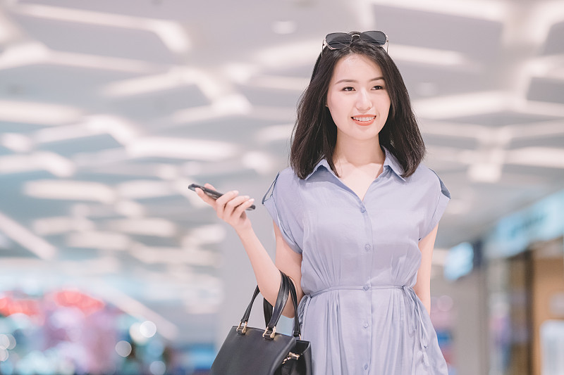一名亚裔华人女性走在购物中心购物，手里拿着智能手机和钱包图片下载