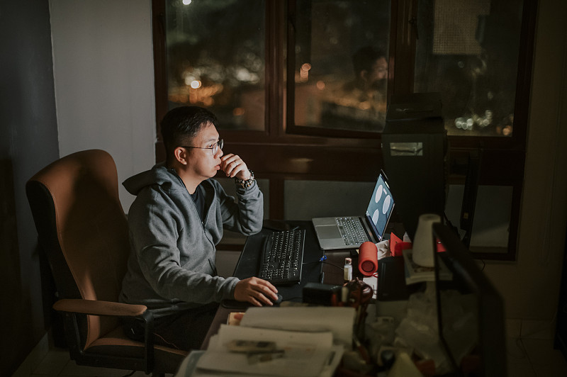 an asian chinese mid adult working at home in his study room alone with hooded shirt in front of desktop PC图片素材