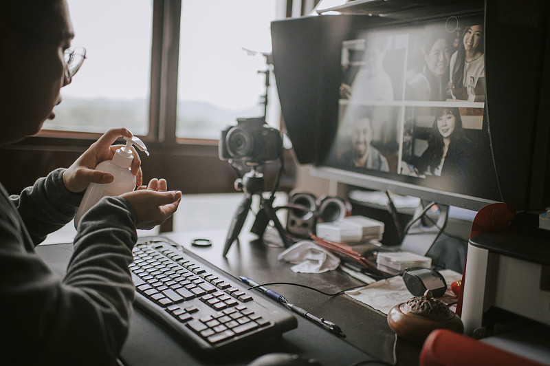 一位亚洲华人男性在他的家庭办公室里做视频/在线博客，用电脑桌面电脑、键盘、鼠标、显示器、蓝牙音箱、带三脚架的单反相机在窗边教授在线摄影课图片下载