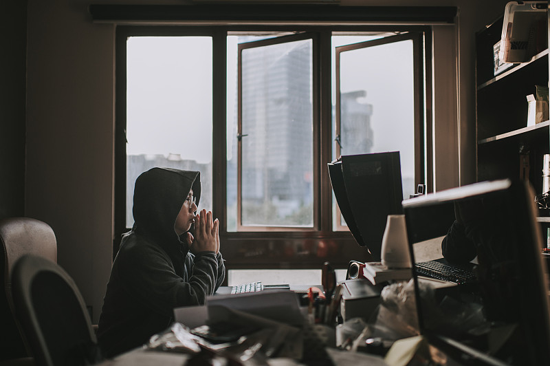 an asian chinese mid adult with hooded shirt looking at computer monitor in his home office during midday with serious facial expression in front of his desk图片素材