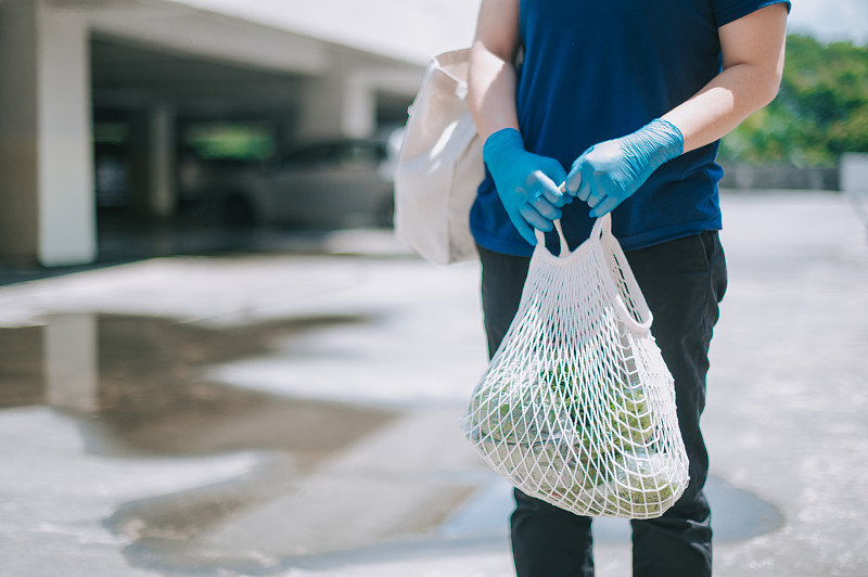 一位亚裔华人中年妇女带着从超市买的食品杂货走在停车场里，戴着医用橡胶手套图片素材