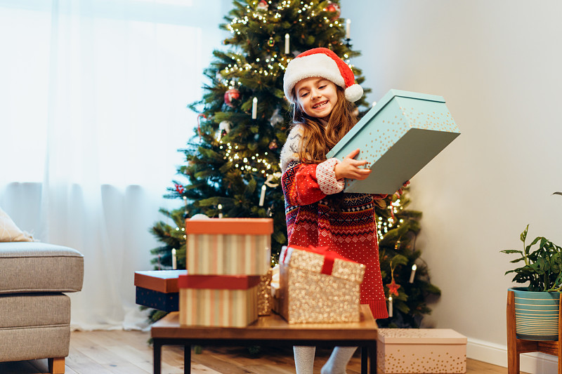 一个快乐微笑的小女孩戴着圣诞帽，穿着喜庆的毛衣，拿着圣诞树背景上的礼品盒。圣诞气氛的概念。准备或打开礼物。有选择性的重点。图片下载