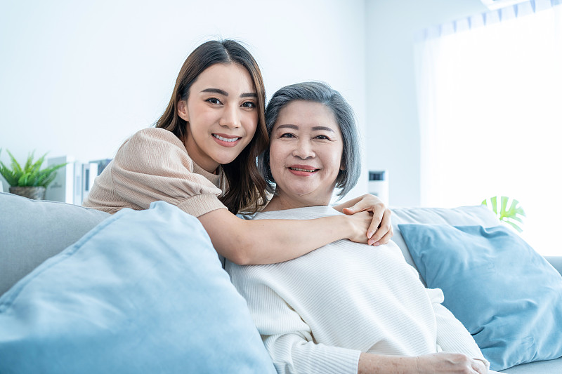 亚洲可爱家庭的肖像，年轻的女儿拥抱年长的母亲。漂亮的女人和年长的妈妈坐在客厅的沙发上，一起在家里享受活动和微笑，看着镜头图片下载