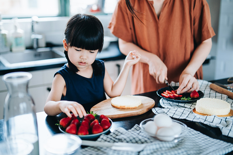 年轻的亚洲母亲和小女儿一起在厨房里烤蛋糕，用草莓和鲜奶油装饰。家庭烘焙。DIY。生日和庆祝概念图片下载
