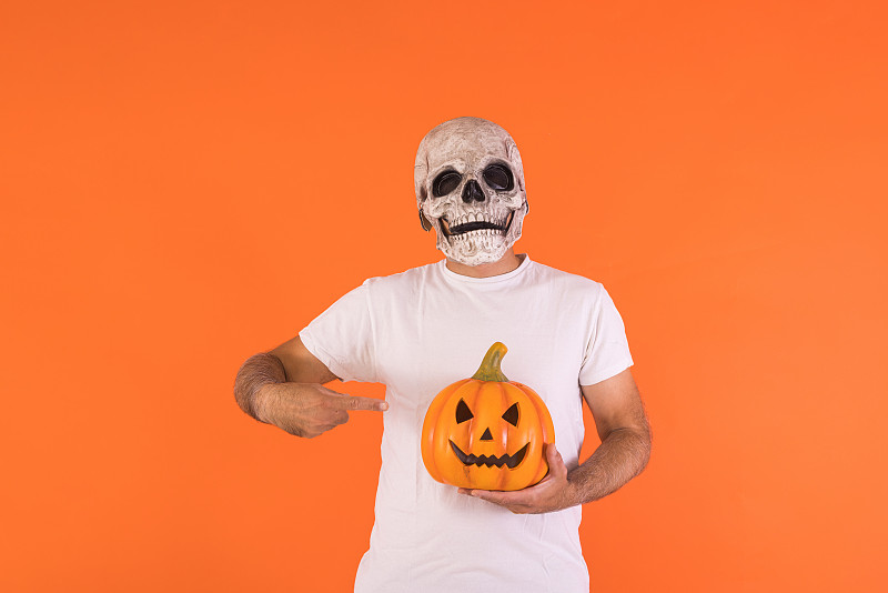 戴着骷髅面具,穿着白色t恤的男子,指着一个有复制空间的南瓜,在橙色