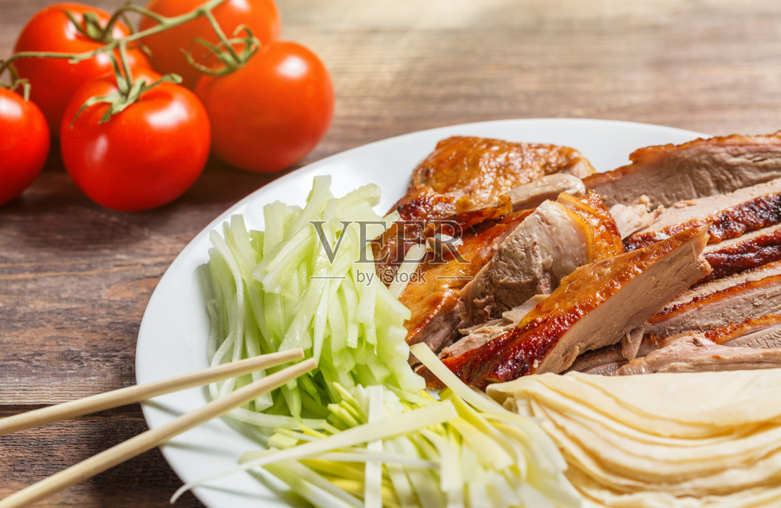 北京烤鸭配洋葱、煎饼、黄瓜片和西红柿放在白色的盘子里，用筷子夹紧。木制的桌子照片摄影图片