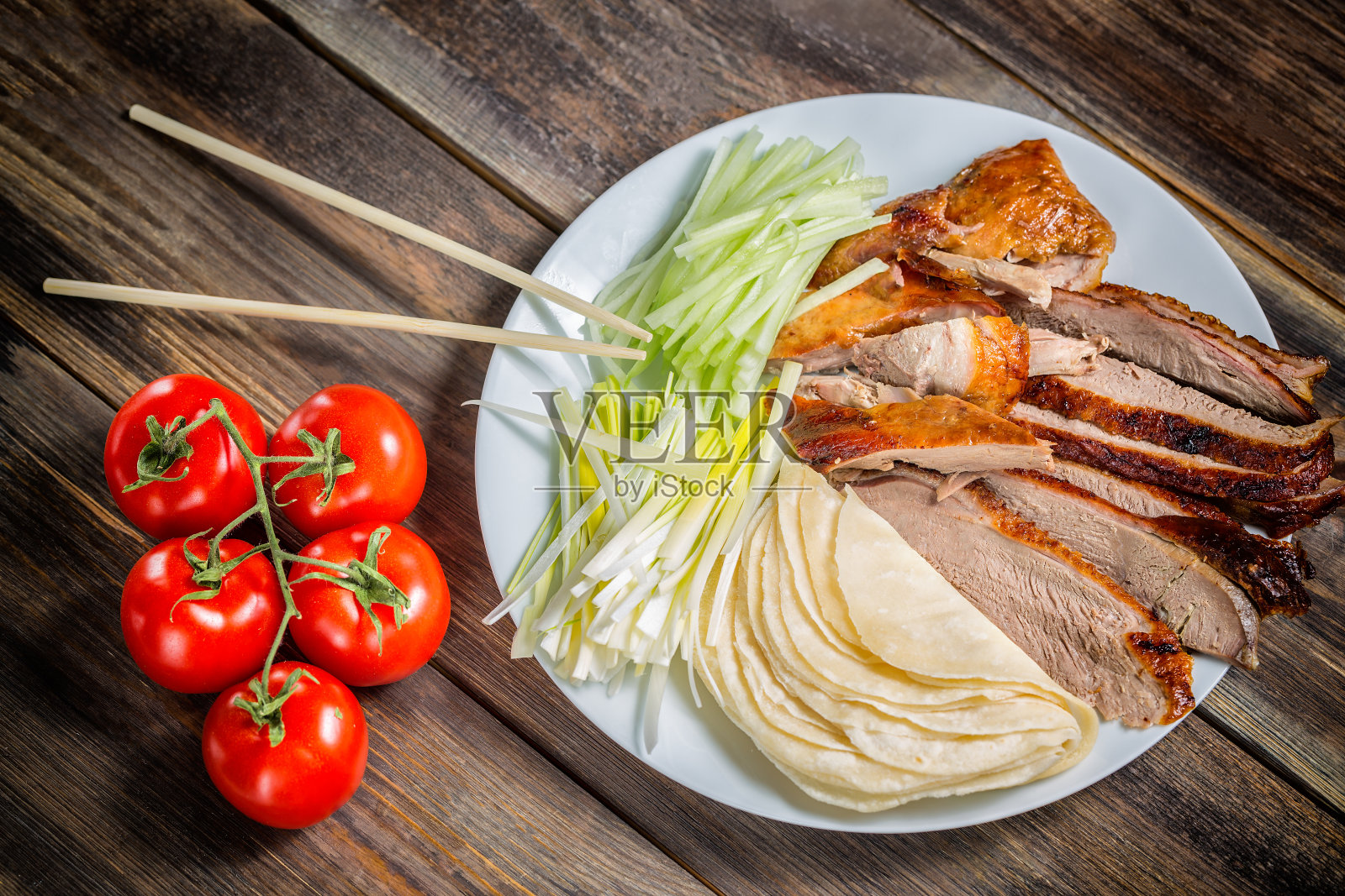 北京烤鸭配洋葱、煎饼、黄瓜片和西红柿放在白色的盘子里，用筷子夹紧。木制桌子和俯视图照片摄影图片