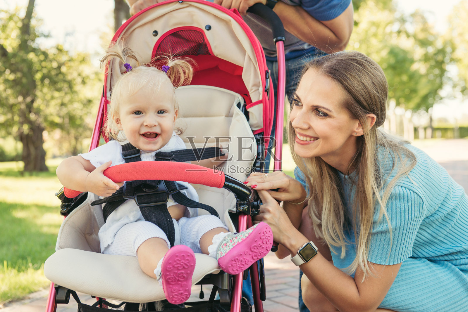 年轻的妈妈和穿着粉色婴儿车微笑的婴儿说话。父母在户外散步的孩子在夏季婴儿车。照片摄影图片