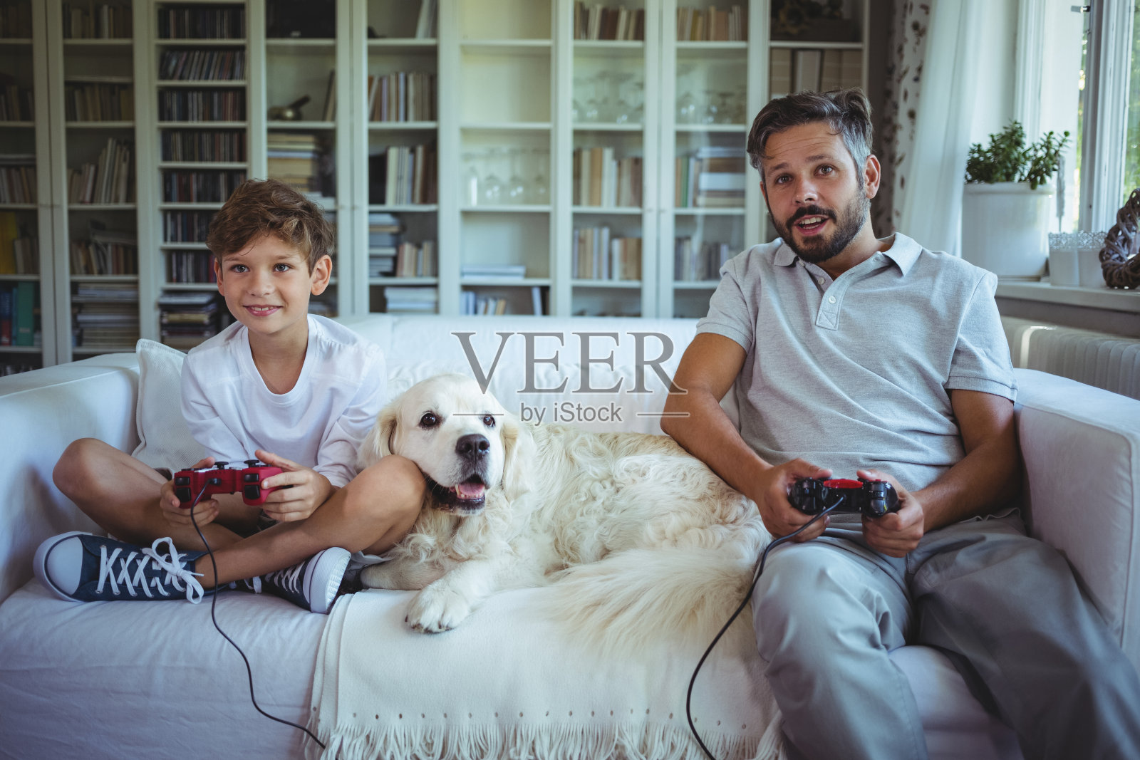 父子俩和宠物狗坐在沙发上玩电子游戏照片摄影图片