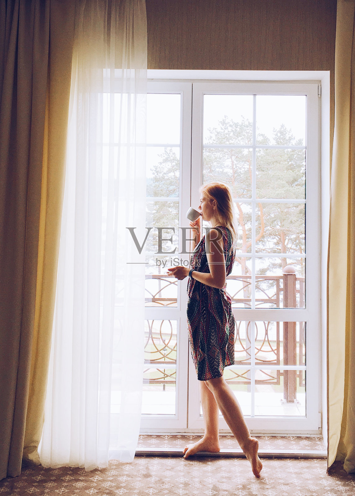 年轻的金发女子站在窗边，用杯子喝咖啡或茶。照片摄影图片