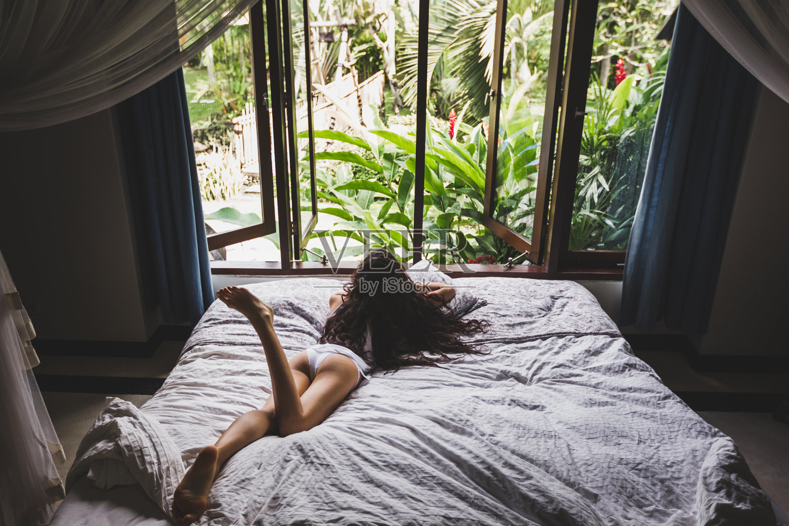 穿着白色内衣的女人懒洋洋地躺在床上，从窗户看热带花园。生活的照片照片摄影图片