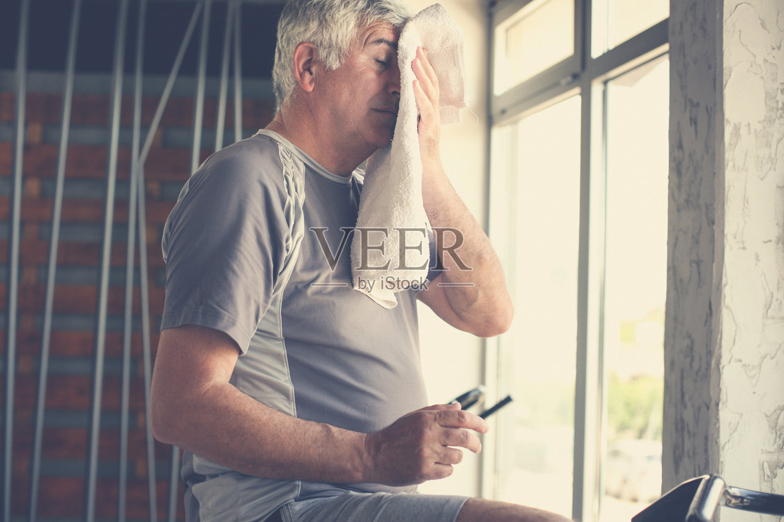 老人坐在健身机上。男子用毛巾擦脸。锻炼在健身房。照片摄影图片