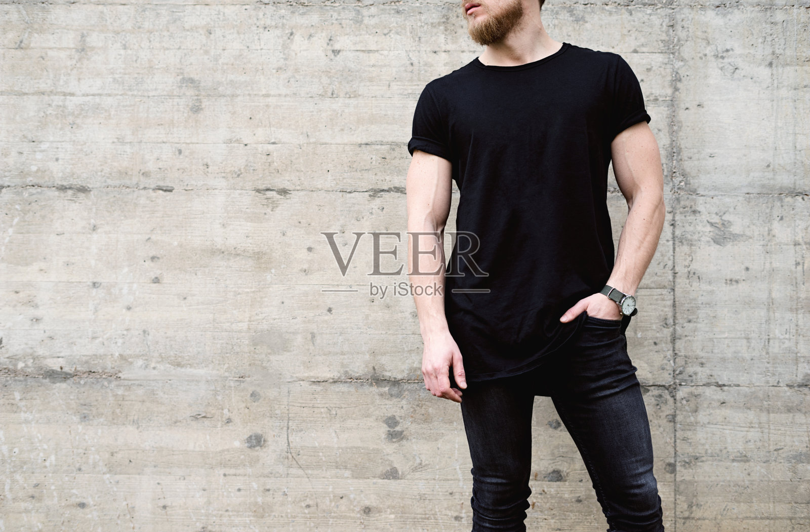 年轻的肌肉胡须男子穿着黑色t恤和牛仔裤在现代城市的中心摆姿势。背景上是空的混凝土墙。Hotizontal模型。照片摄影图片