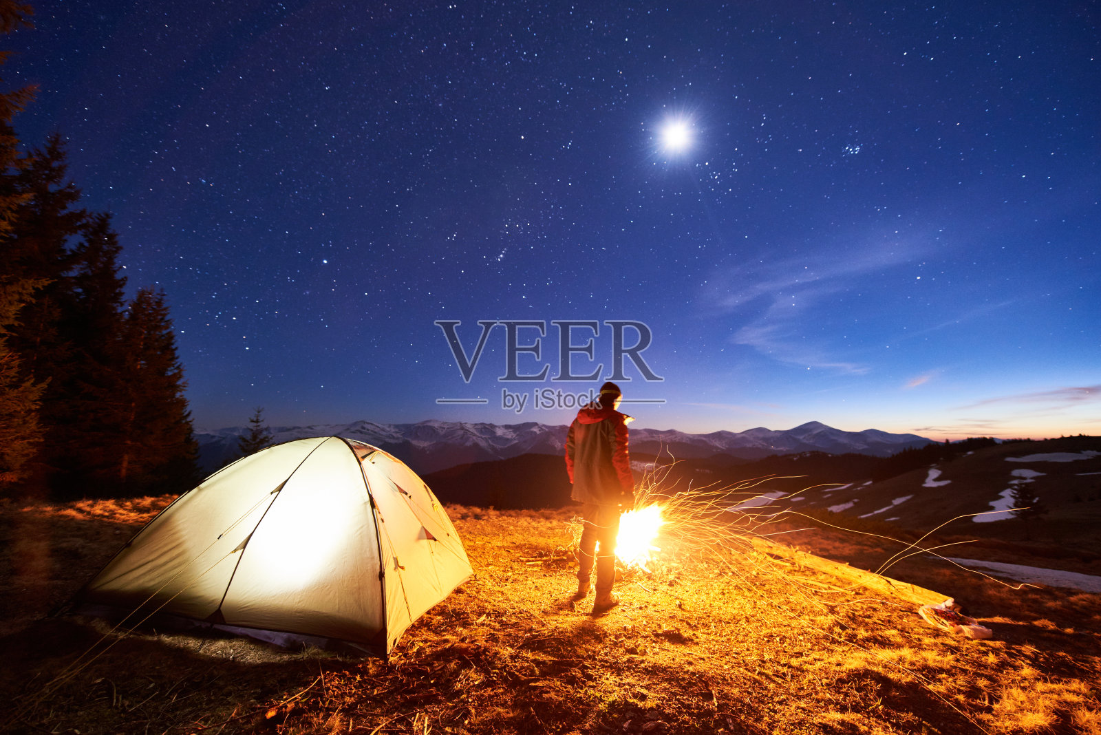 男游客在他的营地休息，站在篝火和帐篷旁边，在美丽的夜空下，满是星星和月亮，欣赏夜景在山里照片摄影图片