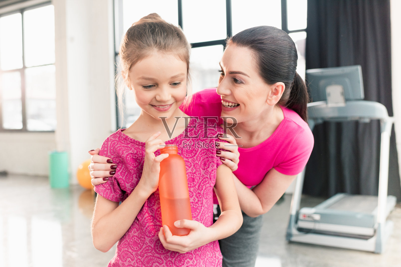 快乐的母亲看着微笑的女儿拿着运动瓶与水在健身房照片摄影图片