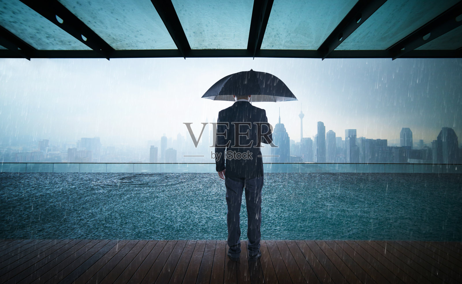 商人撑伞站在天台上，雨天带着城市天际线眺望，风险与危机的概念。照片摄影图片