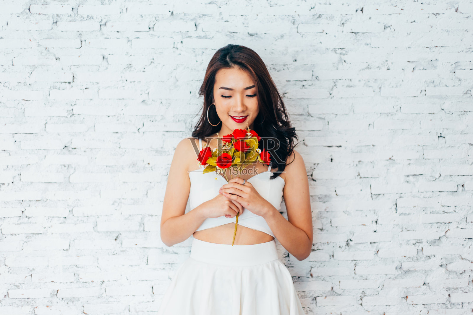 年轻快乐的亚洲女人微笑在时尚的服装和拿着玫瑰花束越过白色的墙的背景照片摄影图片